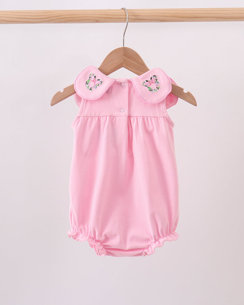 A light pink baby romper with ruffled collar details and embroidered floral accents, hanging on a wooden hanger against a plain light background.