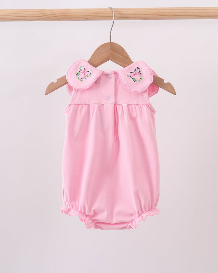 A light pink baby romper with ruffled collar details and embroidered floral accents, hanging on a wooden hanger against a plain light background.
