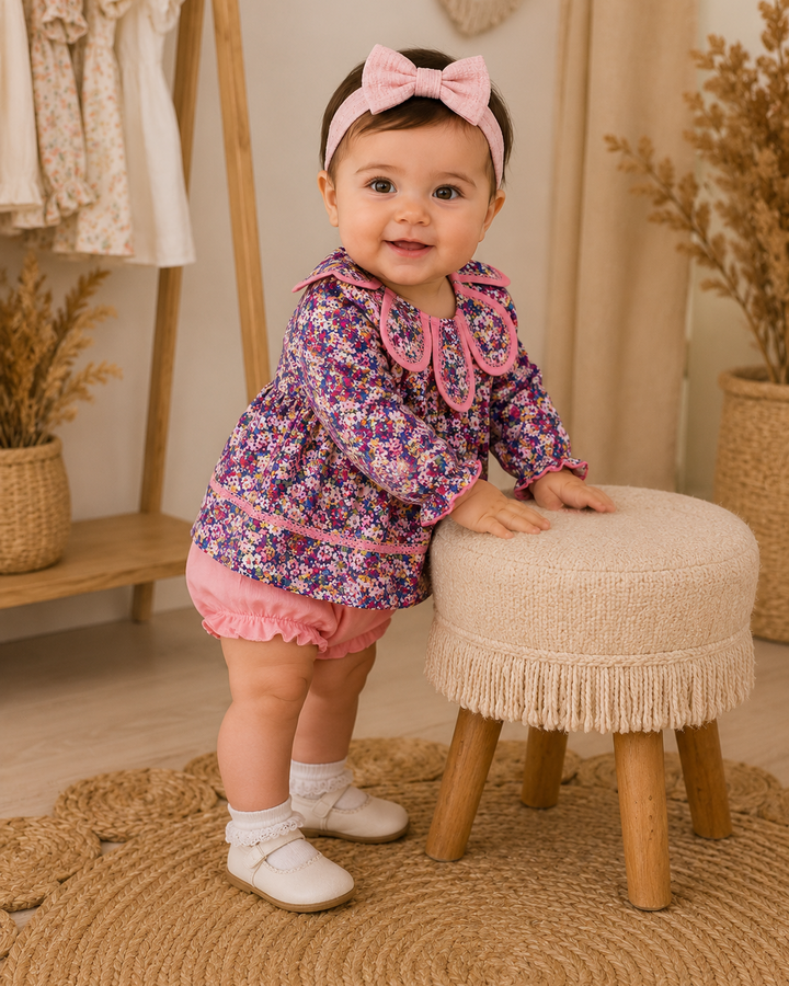 A baby girl wearing a purple and pink floral long-sleeve top with a pink collar, pink ruffled shorts, a pink bow headband, and white shoes, standing next to a small cream-colored stool in a styled nursery setting.