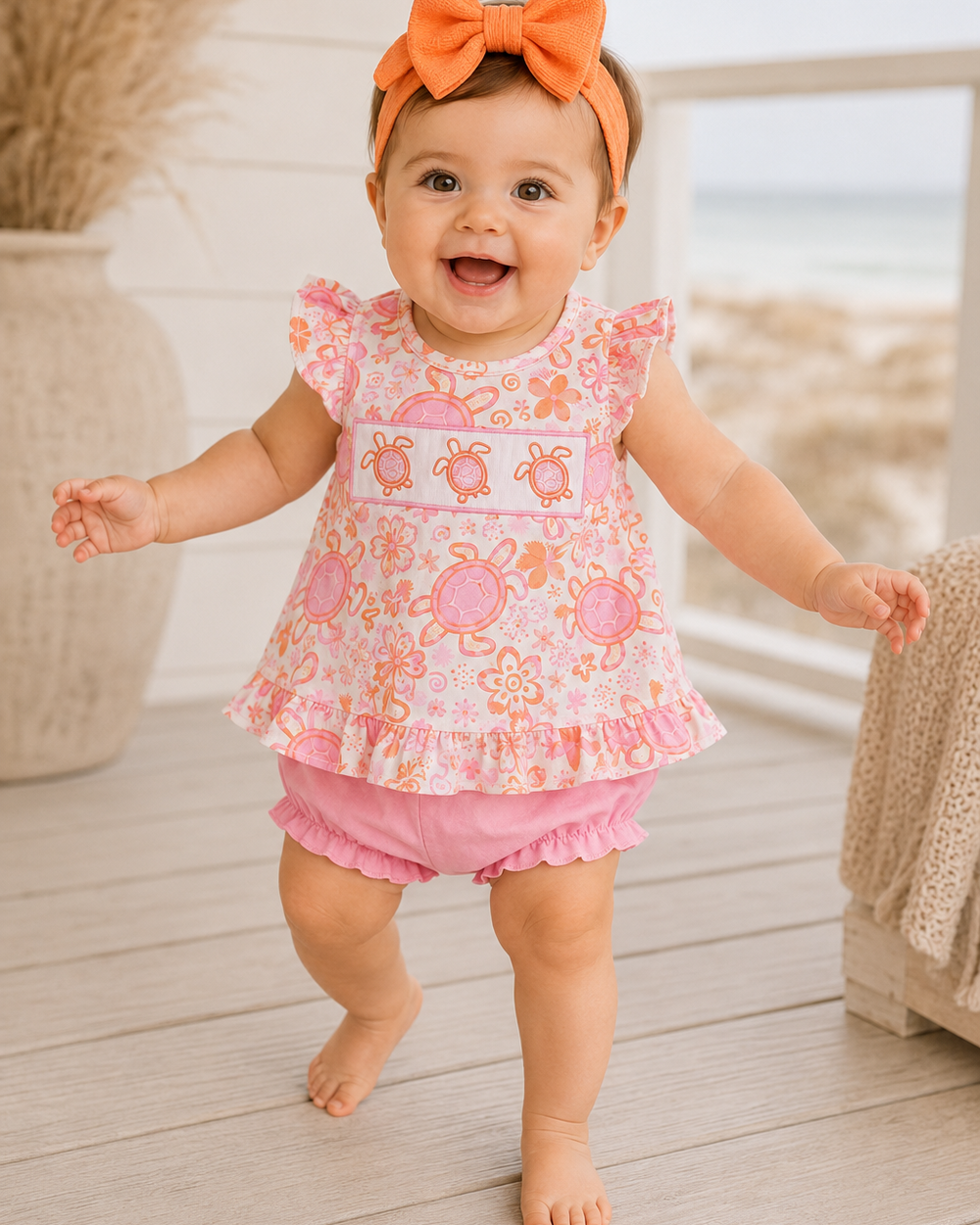Baby in a pink outfit with a floral pattern and orange bow headband standing on a wooden floor.