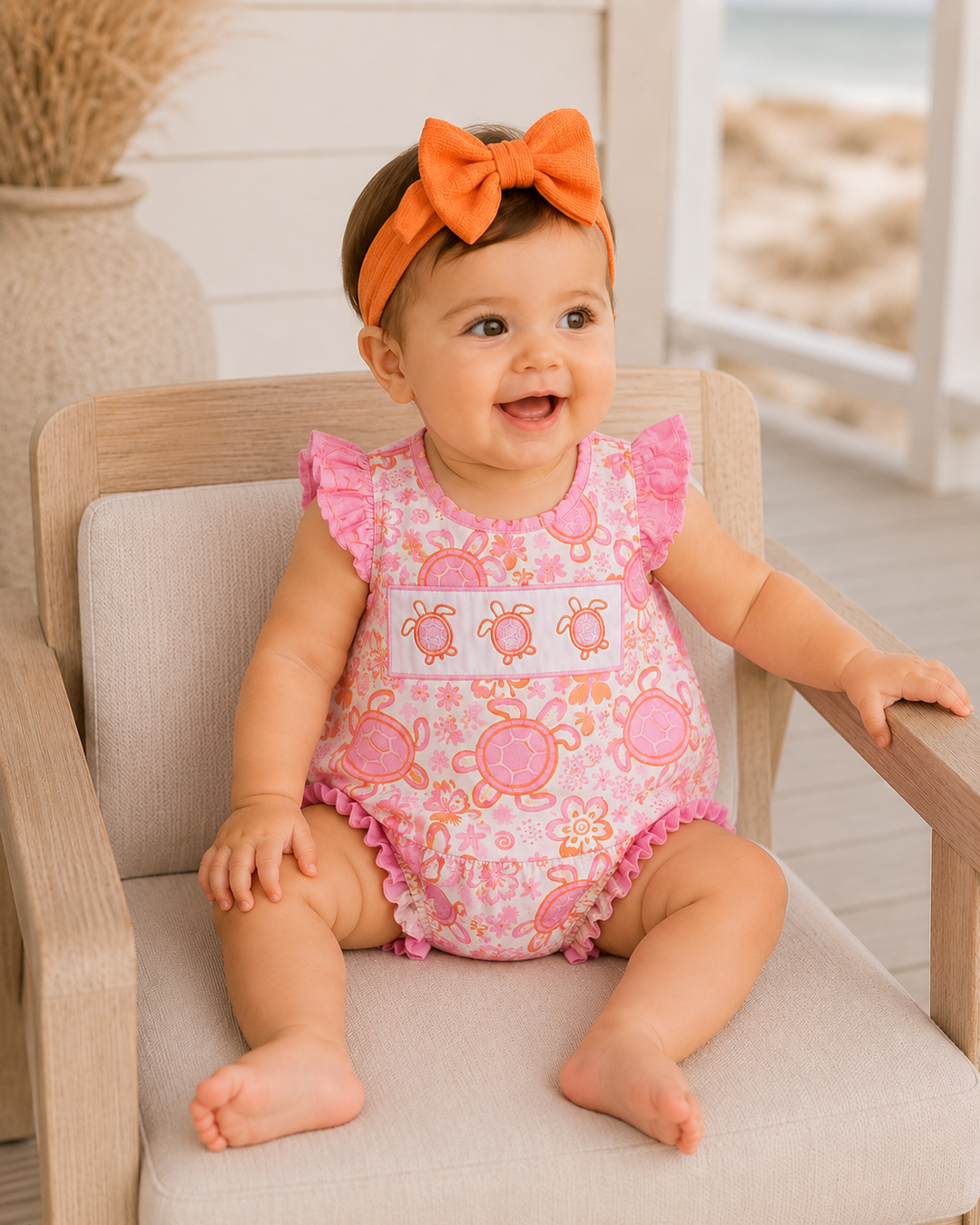 Baby wearing a pink turtle-patterned romper with an orange headband, sitting on a chair.
