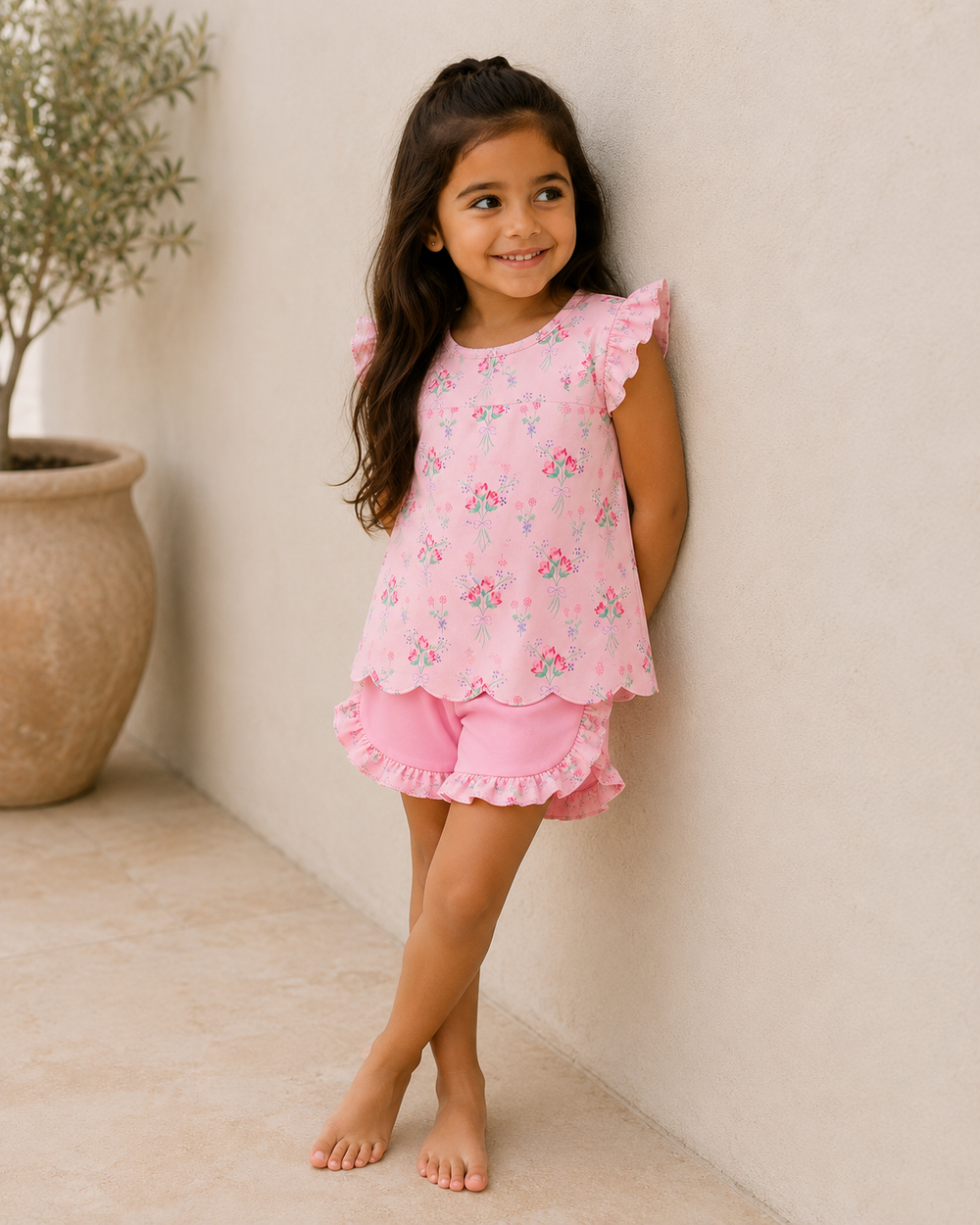 Young girl wearing a pink floral outfit standing against a white wall.
