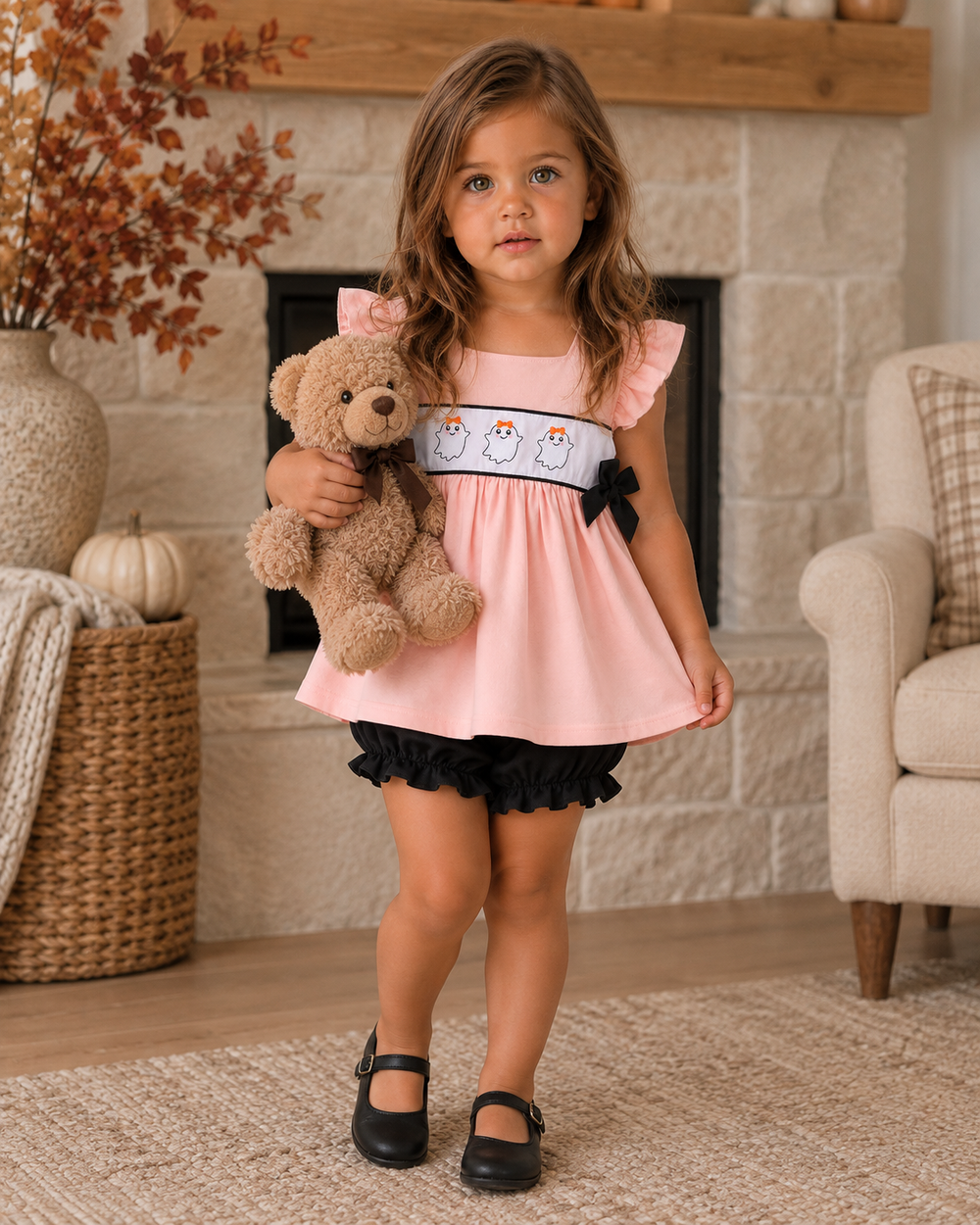 A young girl stands in a cozy living room in front of a stone fireplace, wearing a light pink dress with ruffled sleeves, a white chest band featuring three cartoon penguins, and a black ruffled skirt. She is holding a brown teddy bear and wearing black Mary Jane shoes.