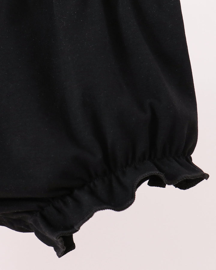Close-up of a black fabric garment with a gathered elastic ruffle hem against a pale pink background.