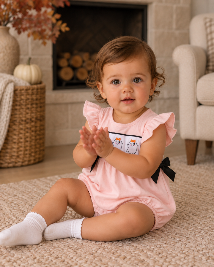 A baby girl sitting on a beige carpet in a cozy living room, wearing a light pink ruffle-sleeve romper with a white chest panel featuring cartoon cats and black ribbon bows.