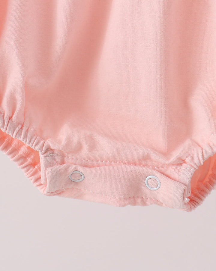 Close-up of a baby romper in light pink fabric, showing the elastic leg openings and two silver snap closures on a plain light background.