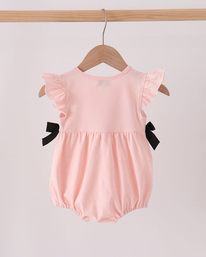 A light pink baby romper with ruffled sleeves and black bow accents on the sides, hanging on a wooden hanger from a wooden clothes rack against a plain light background.