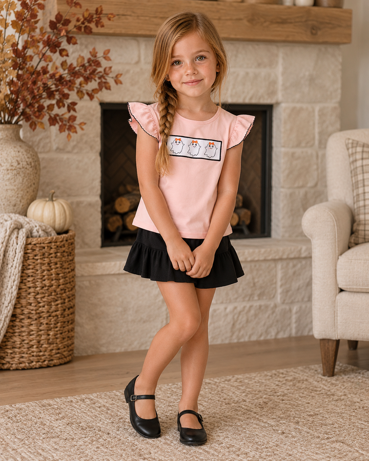 A young girl with a side braid stands in a cozy living room in front of a stone fireplace, wearing a light pink ruffle-sleeve top with a penguin graphic, a black pleated mini skirt, and black Mary Jane shoes.