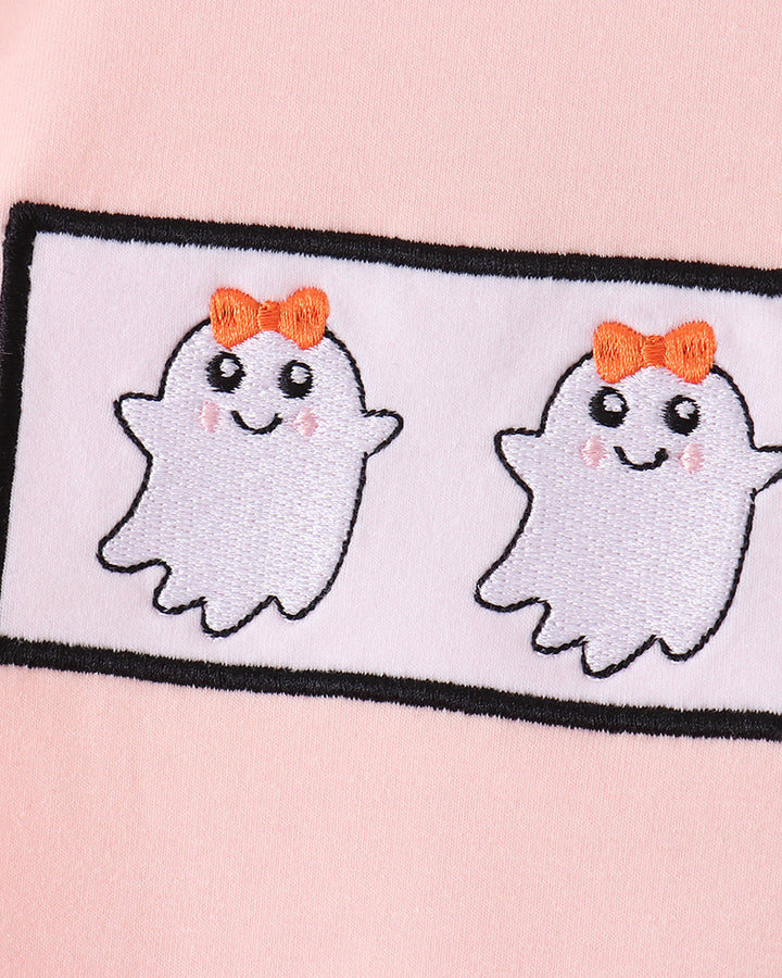 A close-up of a light pink fabric featuring a rectangular embroidered patch with two cute white ghosts wearing orange bows.
