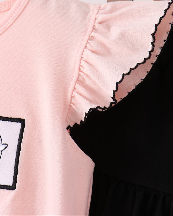 A close-up of a light pink sleeveless top or dress with ruffled shoulders and black scalloped trim, shown hanging on a wooden hanger against a light background. A black garment is partially visible behind it.