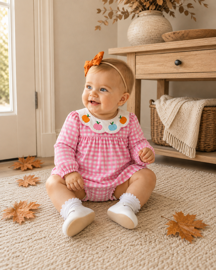 A baby girl sitting on a beige carpet in a cozy room, wearing a pink and white gingham long-sleeve romper with a white bib collar decorated with embroidered fruit, a matching orange bow headband, and white socks.