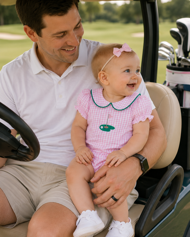 A baby girl in a pink gingham romper with a green Peter Pan collar and a pink bow headband sits on a man&