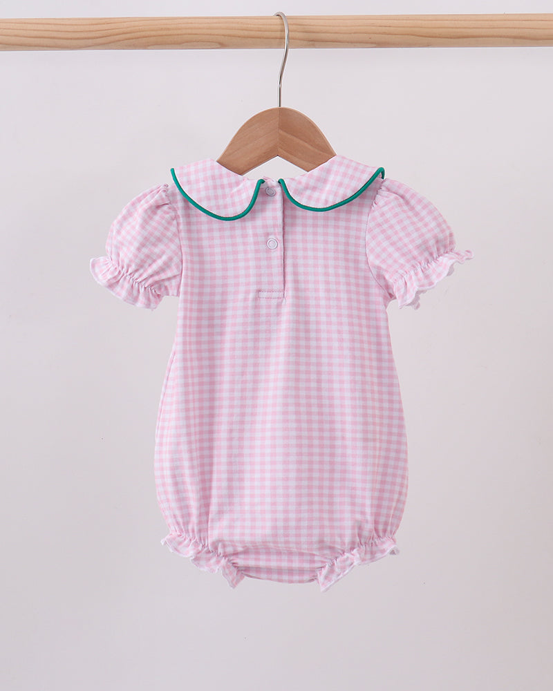 A pink and white gingham baby romper with a green-trimmed Peter Pan collar, hanging on a wooden hanger from a light wood clothing rack against a plain off-white background.