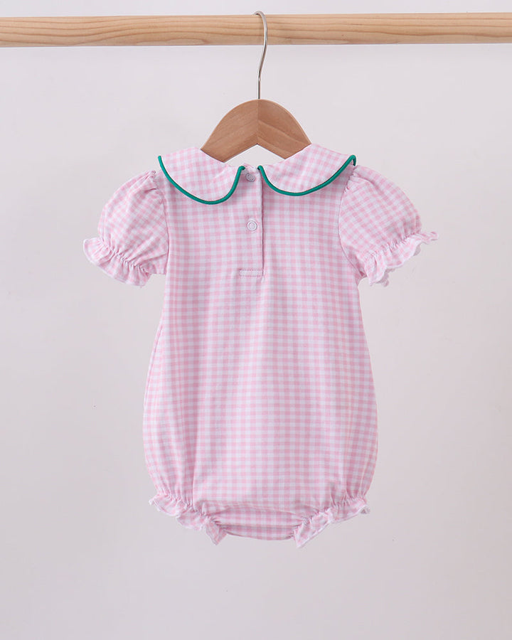 A pink and white gingham baby romper with a green-trimmed Peter Pan collar, hanging on a wooden hanger from a light wood clothing rack against a plain off-white background.