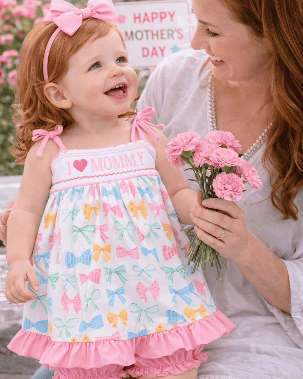 Woman holding a child in a colorful dress with butterfly patterns, both smiling and holding pink flowers on Mother's Day.
