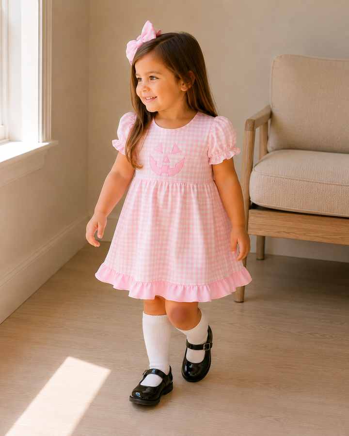 A young girl wearing a pink and white gingham dress with a pink bow headband, white socks, and black Mary Jane shoes, standing in a brightly lit room with a beige armchair in the background.