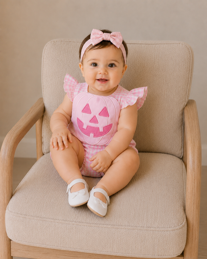 A baby girl sitting on a beige upholstered armchair, wearing a pink and white gingham romper with a pink jack-o&