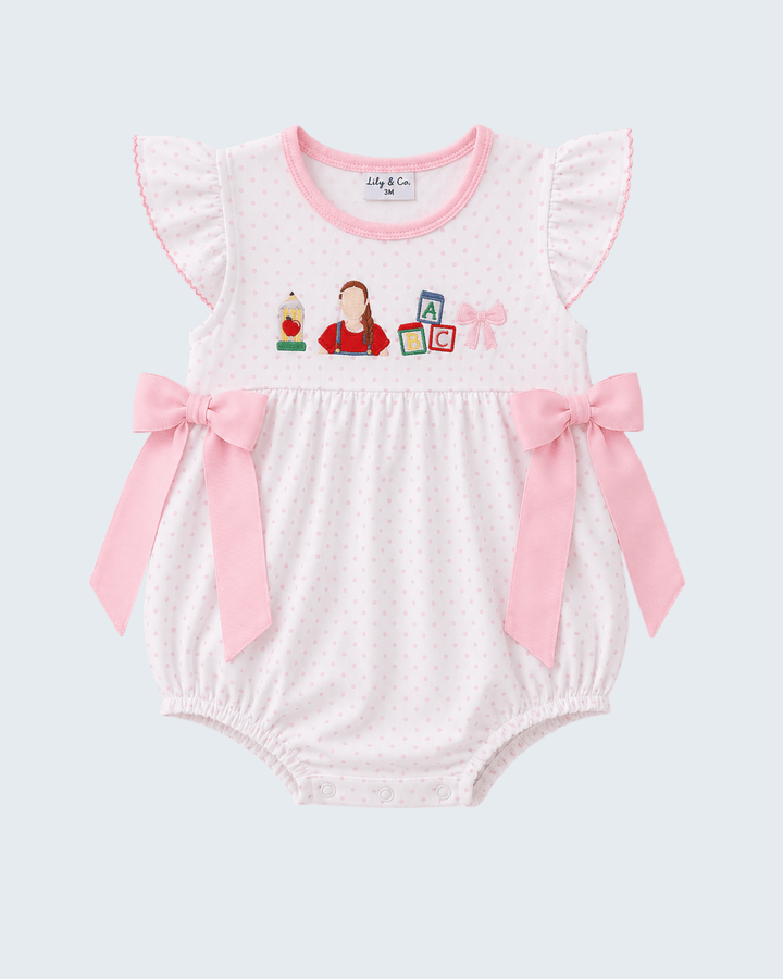 Baby romper in white micro-dot fabric with pink trim, featuring flutter sleeves, pink ribbon bows at the sides, and a colorful cartoon graphic on the chest.