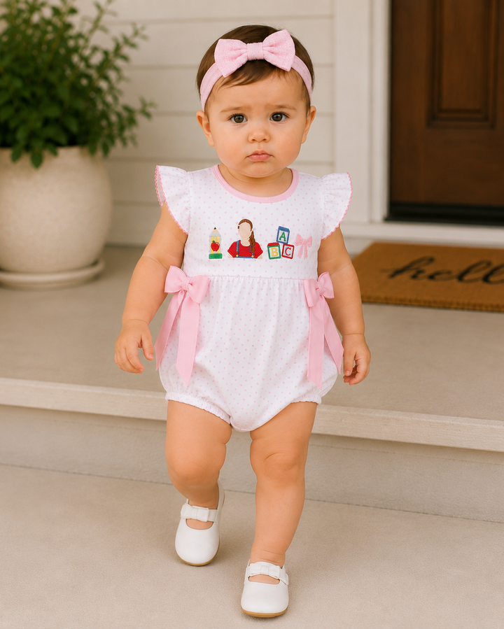 A toddler girl wearing a white romper with pink ruffle sleeves and side bows, a pink bow headband, and white shoes, standing on front steps in front of a house door.
