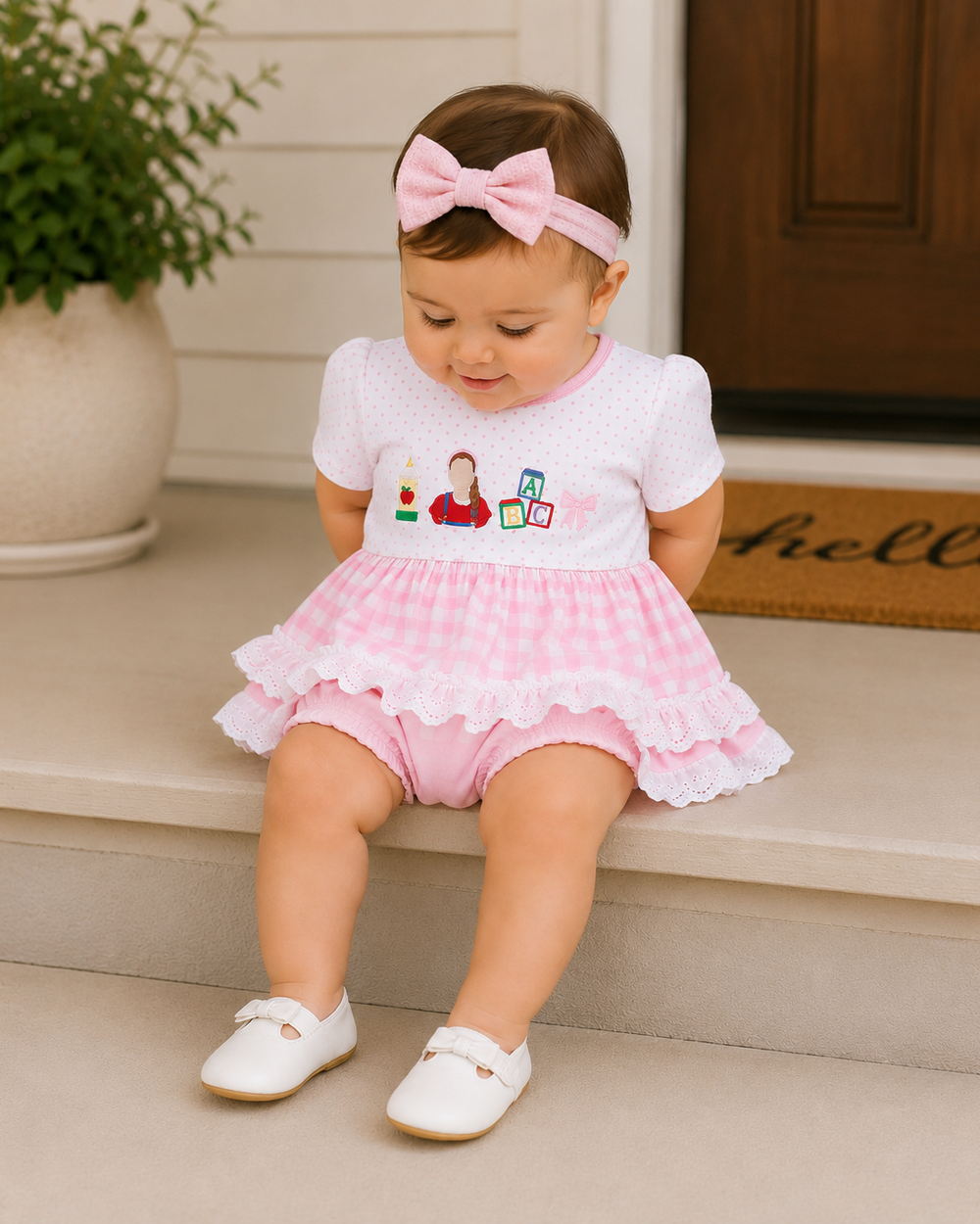A toddler girl sits on outdoor steps wearing a pink and white gingham romper with a ruffled skirt, a matching pink bow headband, and white shoes.