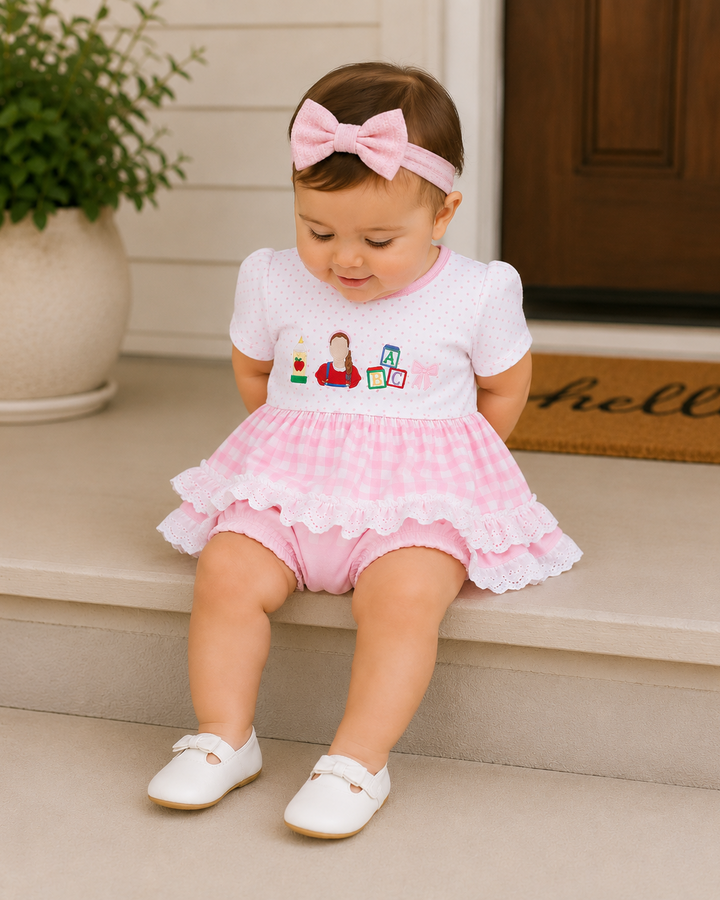 A toddler girl sits on outdoor steps wearing a pink and white gingham romper with a ruffled skirt, a matching pink bow headband, and white shoes.