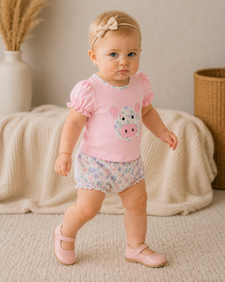 A toddler girl wearing a pink short-sleeve top with a pig face applique and matching floral shorts, standing in a styled indoor room with a knitted blanket, woven basket, and dried grass in a vase.