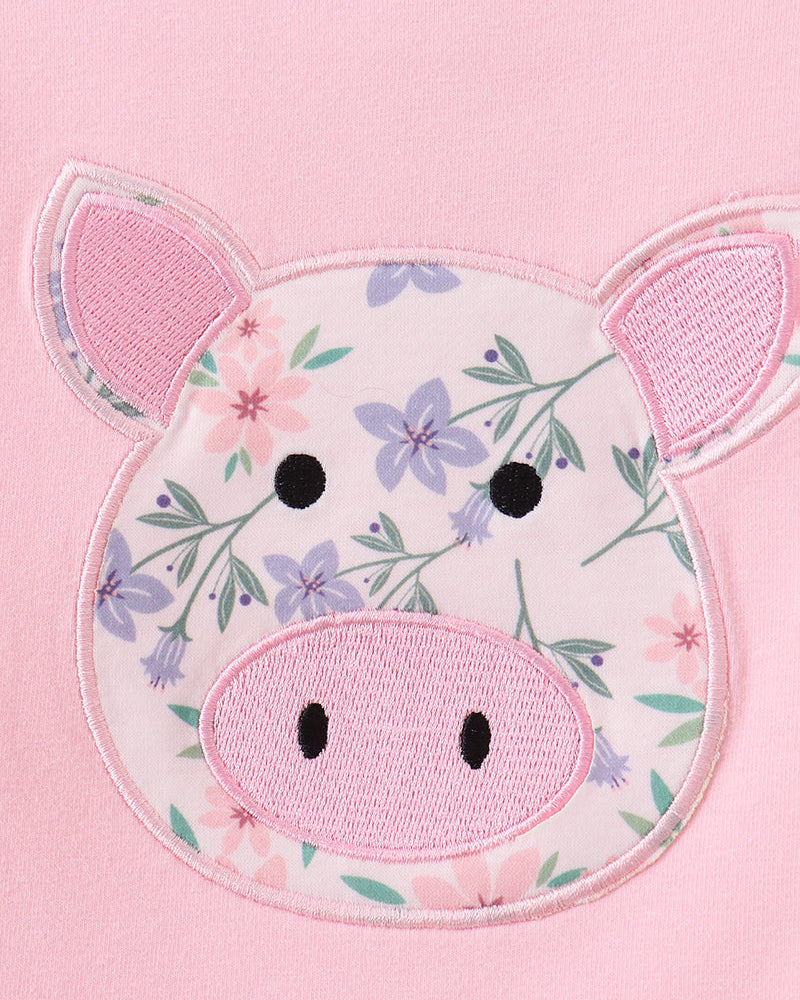 A close-up of a pink garment featuring a large applique pig face patch with a floral pattern, pink snout, and pink ears.