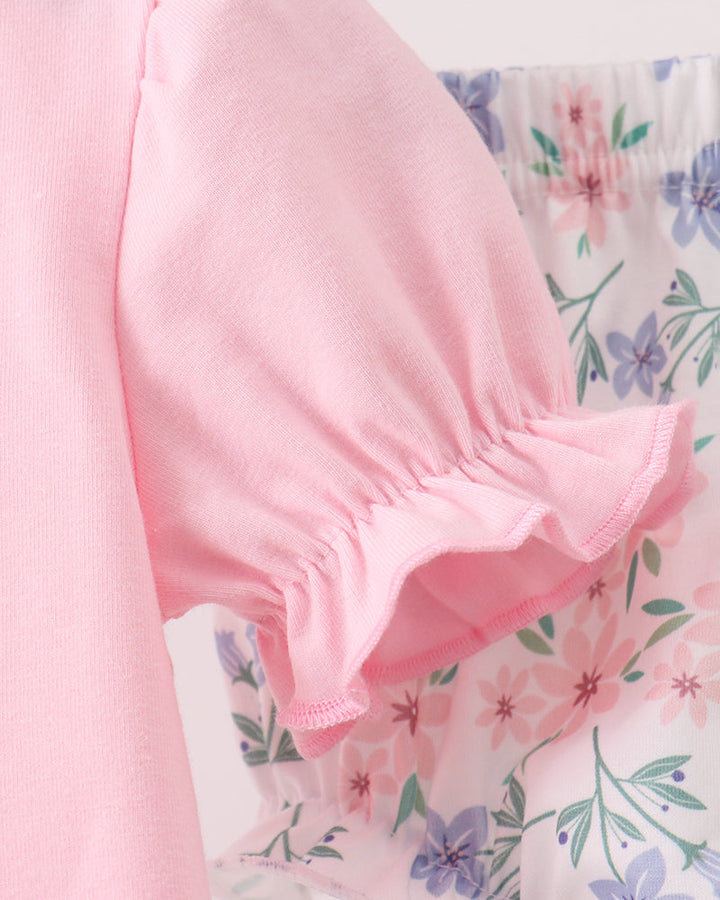 Close-up of a light pink short-sleeve top with a ruffled hem, shown hanging next to a white floral skirt or shorts.