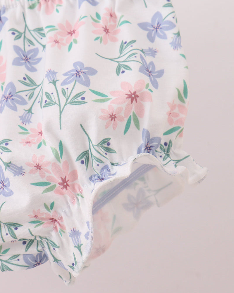 Close-up of a white fabric garment with a pastel floral print featuring pink and blue flowers with green stems, showing a ruffled hem against a pale pink background.