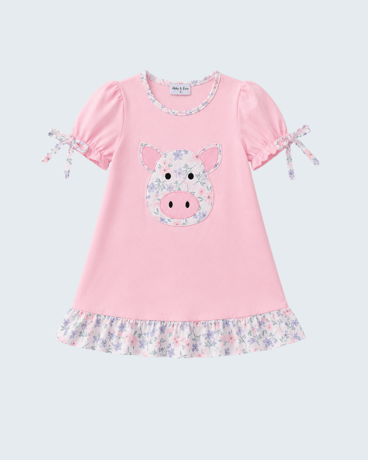A pink short-sleeve baby dress with a floral pig face applique on the front, ruffled trim at the collar and hem, and tie details on the sleeves, shown against a light gray background.
