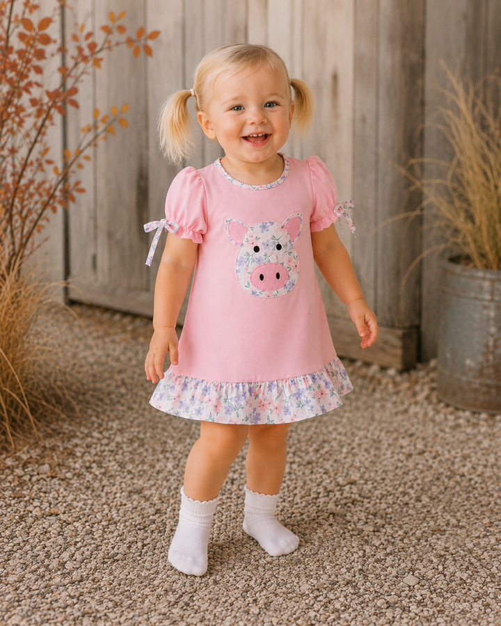 A young toddler girl with blonde pigtails stands outdoors wearing a light pink short-sleeve dress with a cow applique on the chest, a floral ruffled hem, and white socks. The background features a weathered wooden fence, gravel ground, and potted plants.