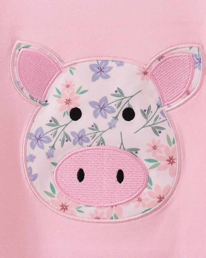 A close-up of a pink garment featuring a pig face applique with a floral print on the forehead, pink embroidered ears, and black embroidered eyes and nostrils.