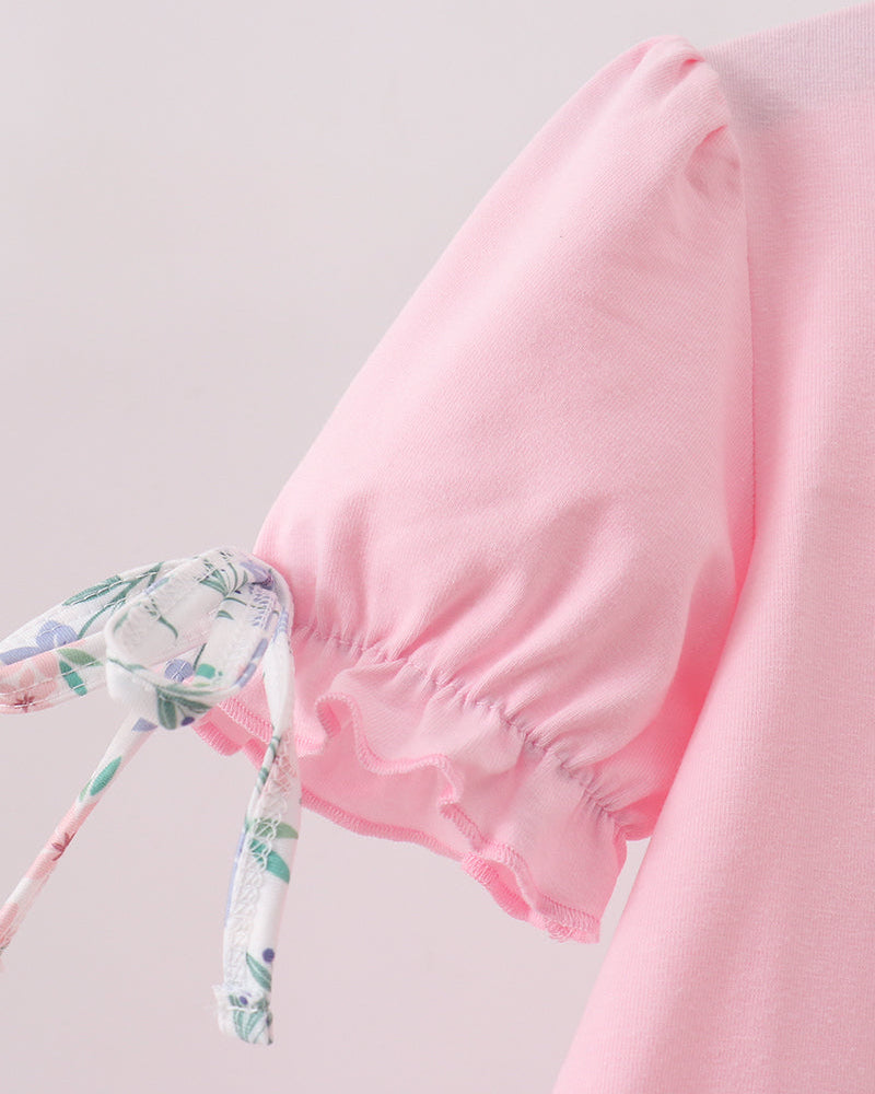 A close-up of a light pink garment featuring a gathered puff sleeve with a ruffled hem and a matching floral print tie detail.