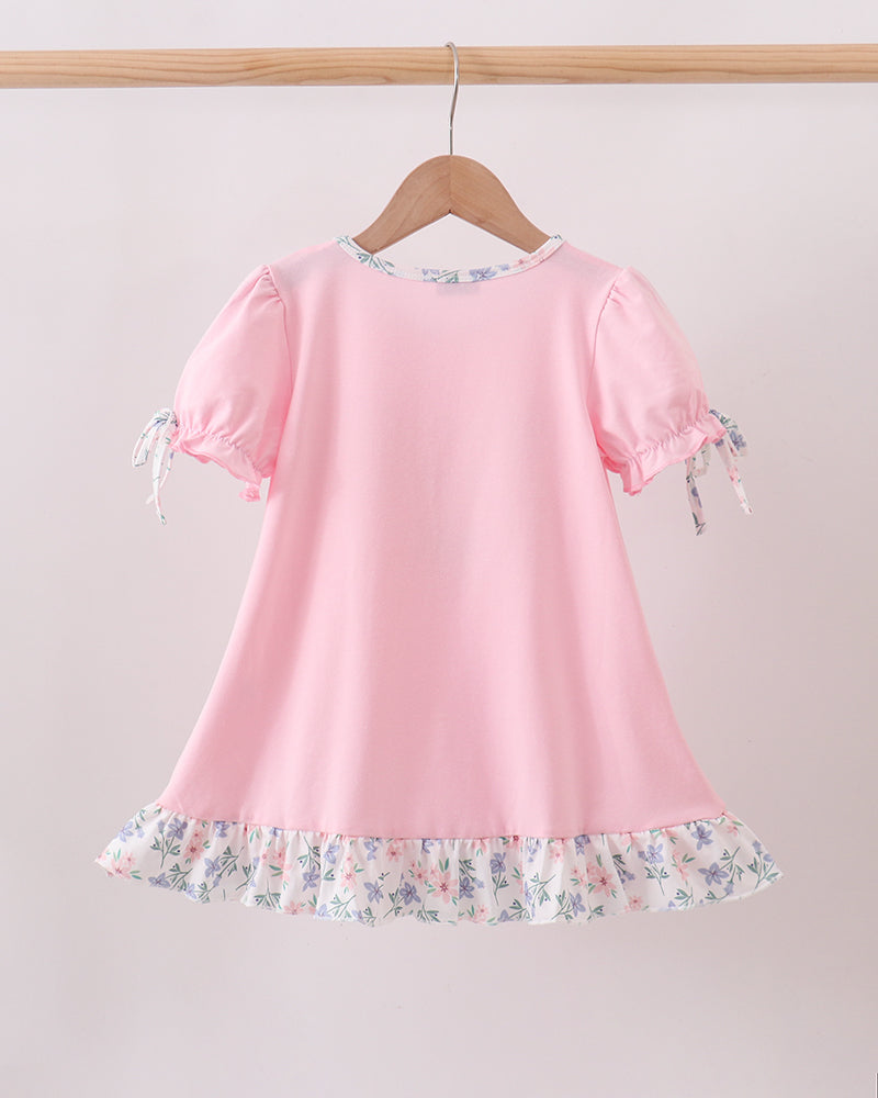 A pink short-sleeve dress with a ruffled floral hem and matching tie details on the sleeves, hanging on a wooden hanger against a light pink background.