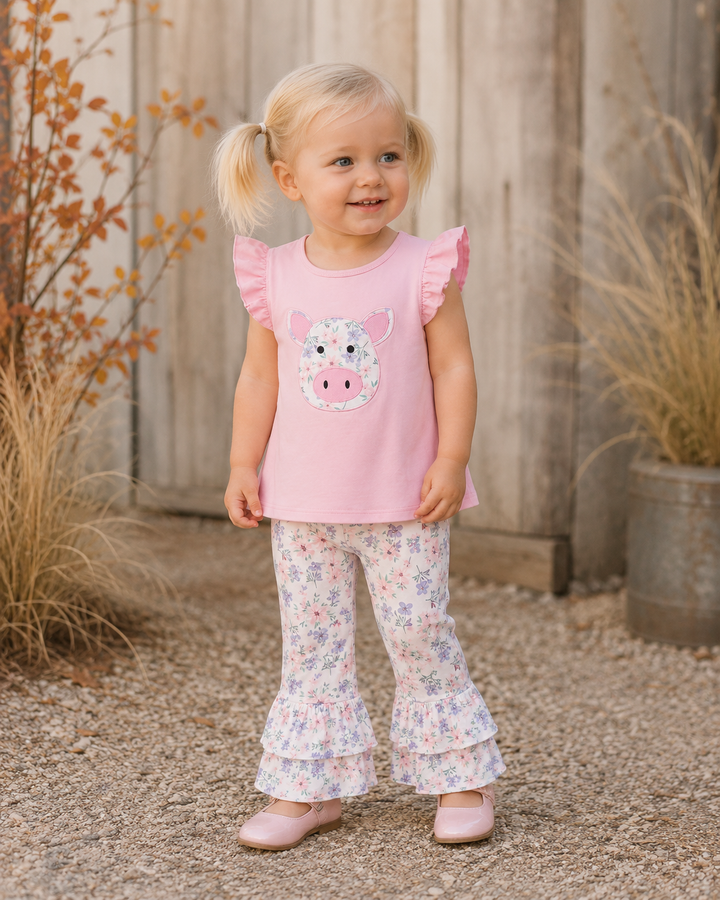 A young girl with blonde pigtails stands outdoors in front of a weathered wooden fence, wearing a light pink sleeveless top with a pig face applique and ruffled shoulders, paired with white ruffled flare pants covered in a small floral print, and pink shoes.