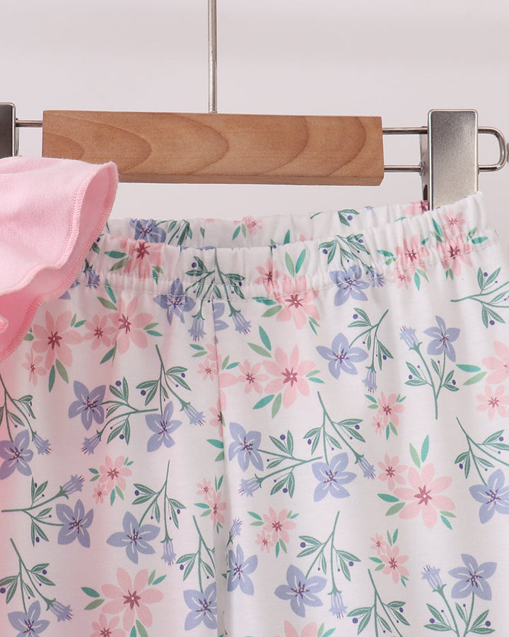 A close-up of floral pajama shorts or a matching set hanging on a wooden hanger. The fabric is white with an all-over print of pink and purple flowers with green leaves, and a soft pink fabric is visible on the left edge.