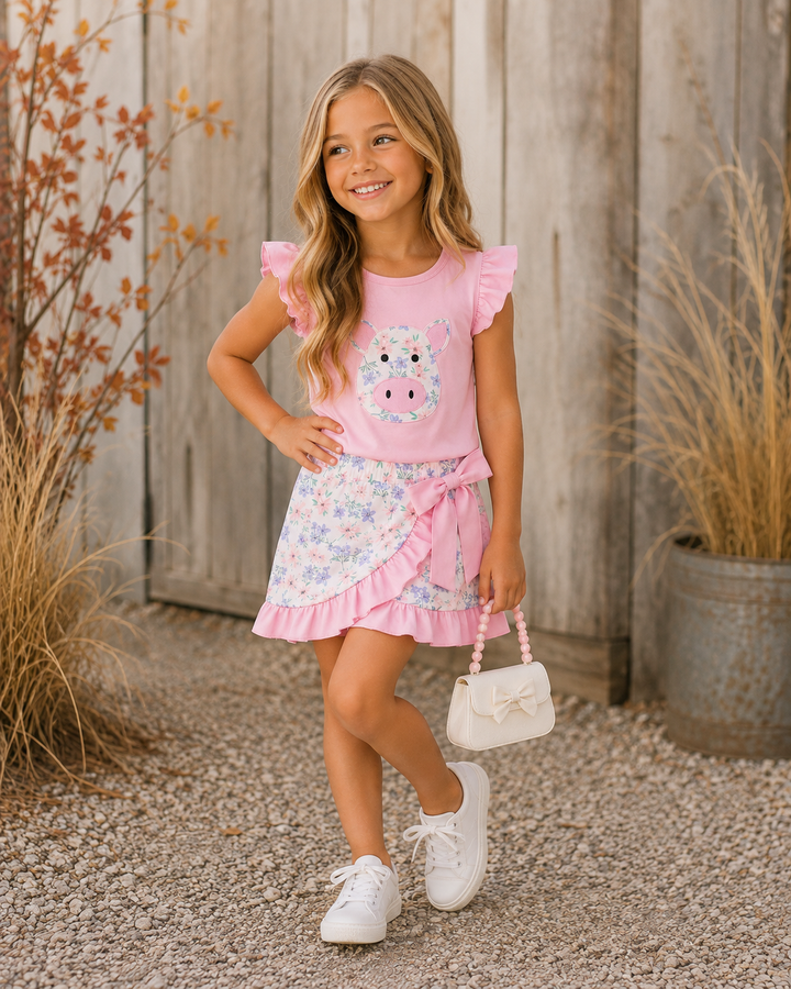 A young girl stands outdoors in front of a weathered wood fence, wearing a pink ruffle-sleeve top with a cow graphic and a matching floral ruffled skirt. She is holding a small white handbag and wearing white sneakers.