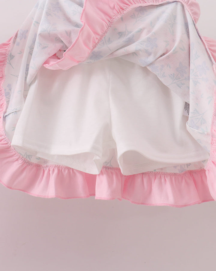 Pink ruffled baby shorts with a white inner lining and a light blue floral print outer layer, shown hanging against a pale pink background.