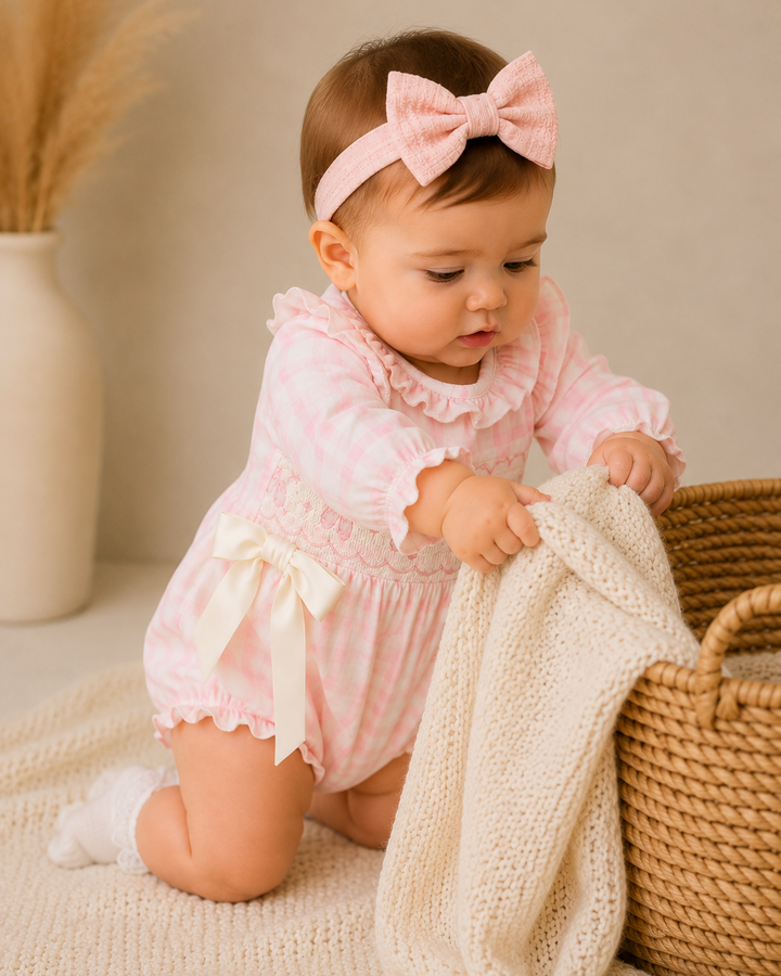 A baby girl wearing a pink and white gingham smocked romper with a matching pink bow headband, kneeling on a cream knit blanket next to a woven basket and holding a cream knit throw.