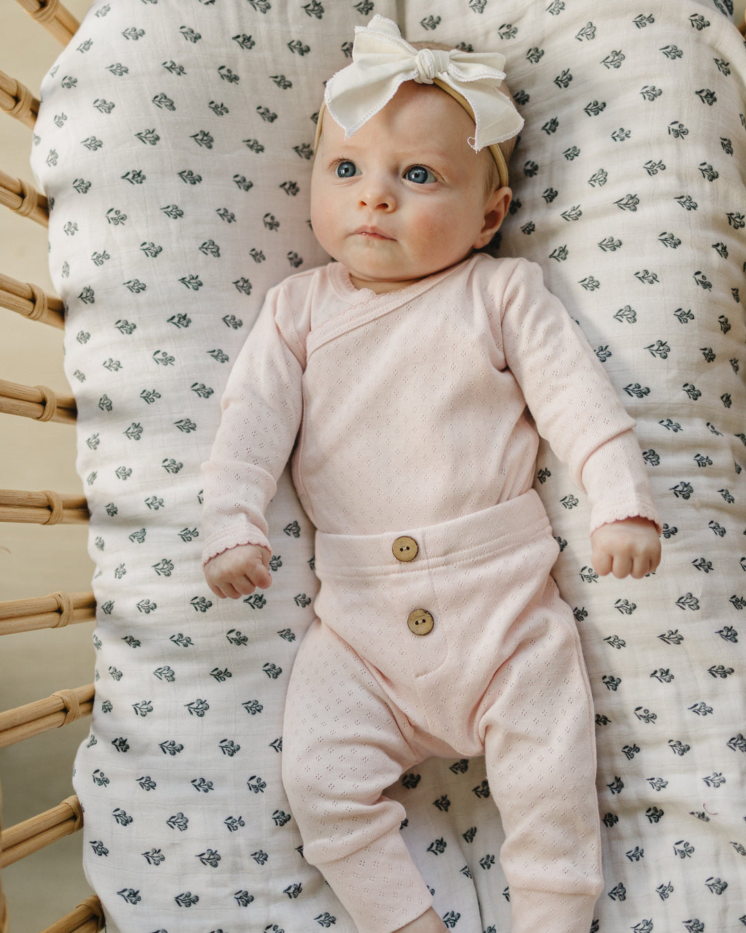 Baby in a pink onesie with buttons, sitting on a textured chair with a patterned cushion.