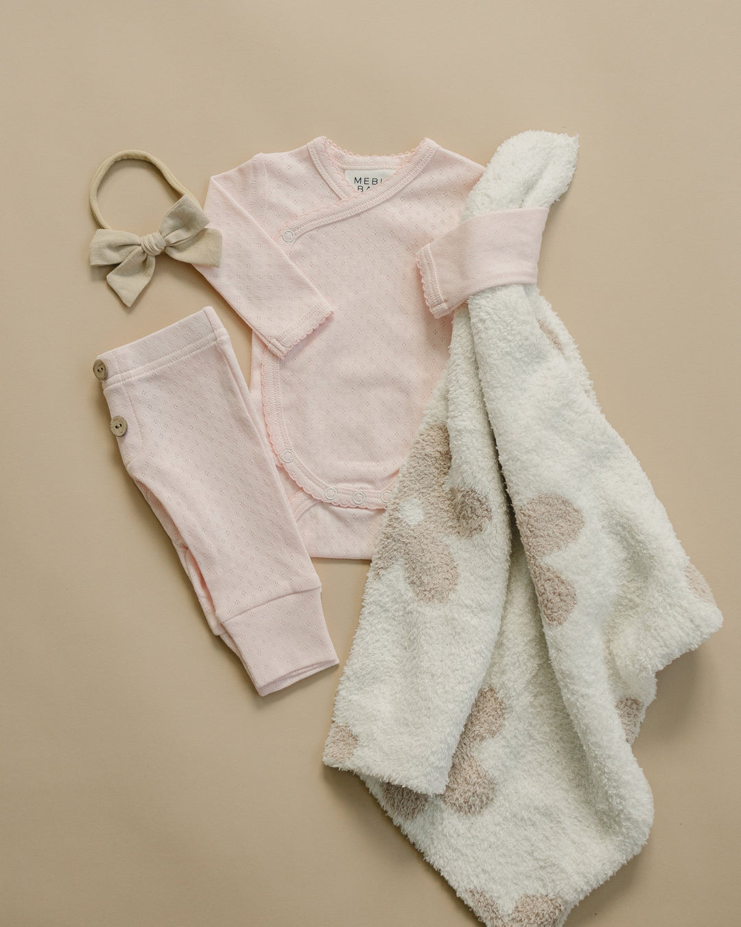 Baby clothing set with pink outfit, pants, blanket, and headband on a beige background