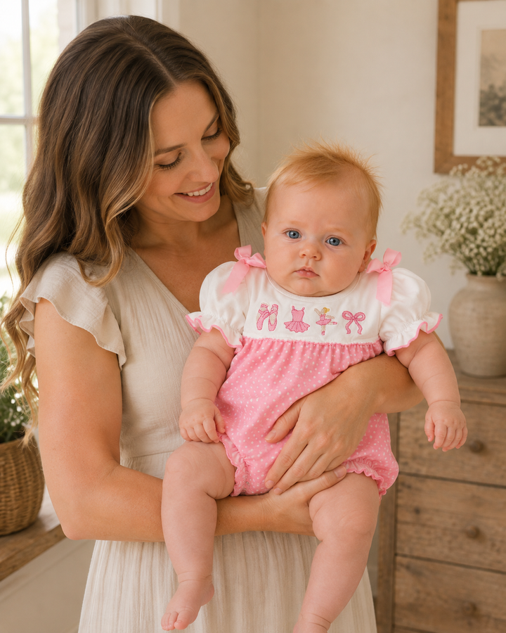 A woman holding a baby wearing a pink and white romper. The romper has a white puff-sleeve top with pink embroidered details and a pink polka-dot skirt.