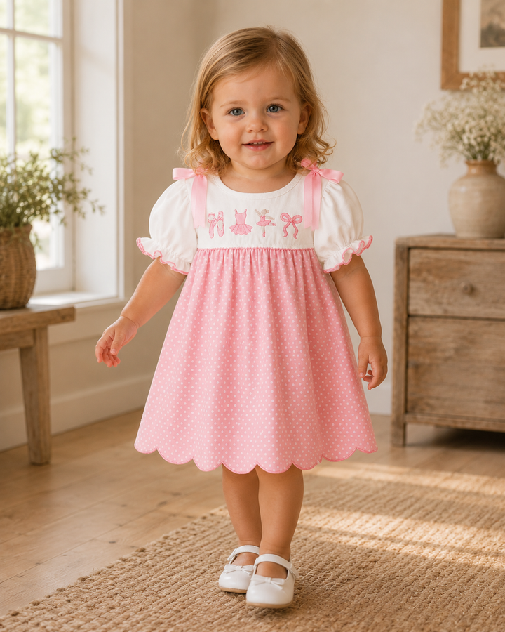 A toddler girl wearing a pink and white dress with white Mary Jane shoes, standing in a bright, sunlit room with wooden floors and a rustic dresser.