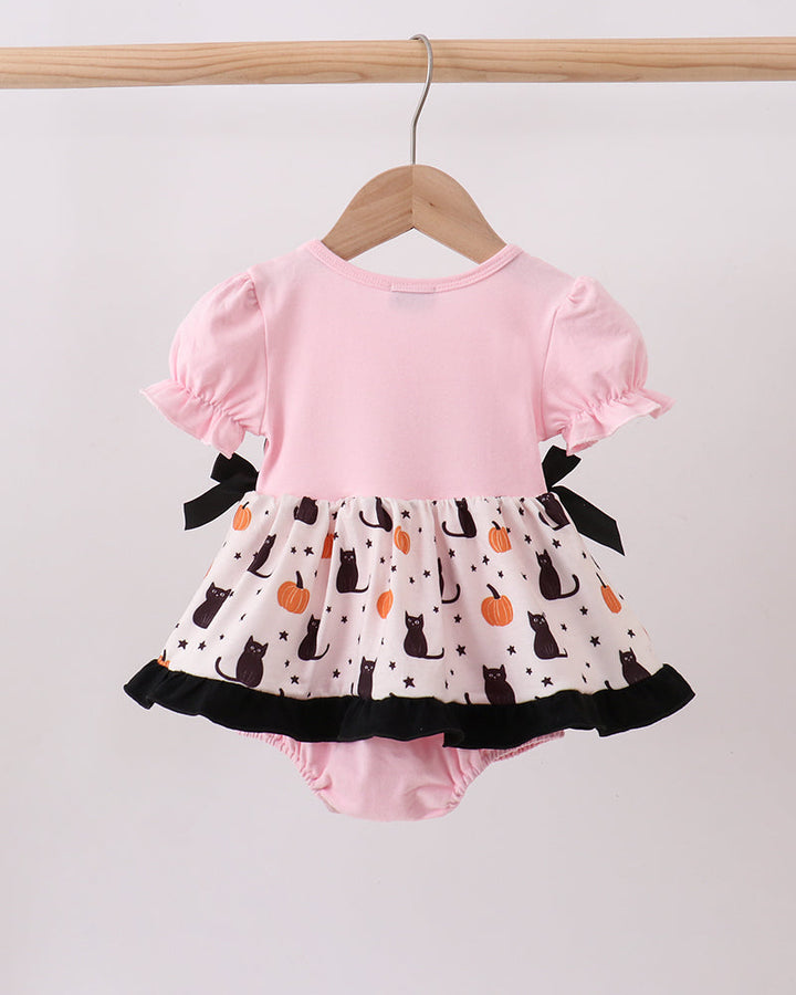 A baby romper with a light pink top and a white ruffled skirt featuring a black cat and orange pumpkin pattern, hanging on a wooden hanger.