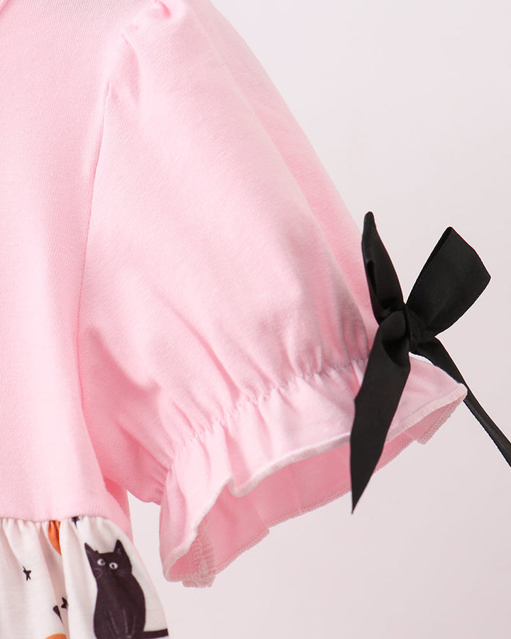 Close-up of a light pink short-sleeve top with a black ribbon bow at the cuff, paired with a white skirt featuring a black cat and orange pumpkin print.