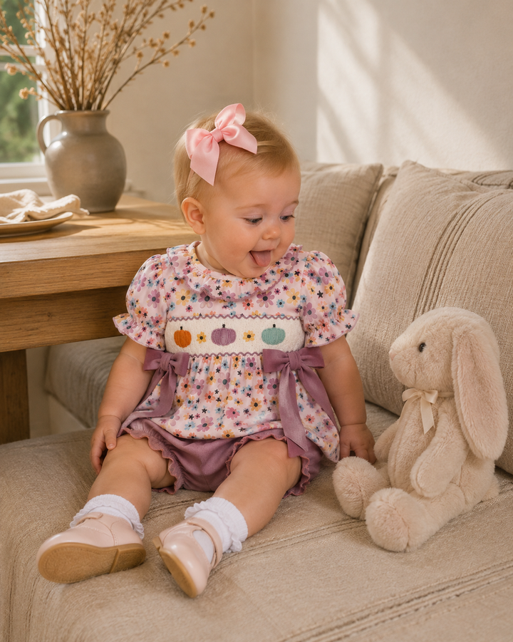 A baby girl sitting on a beige couch next to a plush bunny toy, wearing a floral print romper with purple ruffle trim and a pink bow in her hair.