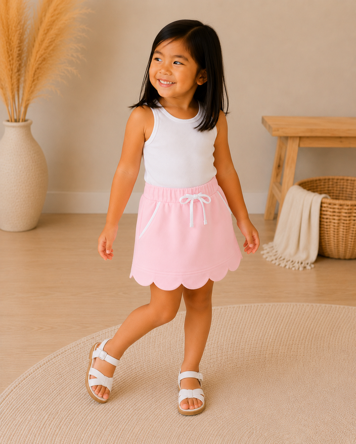 A young girl smiling and walking while wearing a two-piece outfit consisting of a white sleeveless top and a light pink skort with a scalloped hem and white drawstring. She is standing on a round beige rug in a warm, neutral-toned room with a wooden bench, woven basket, and pampas grass in a vase.