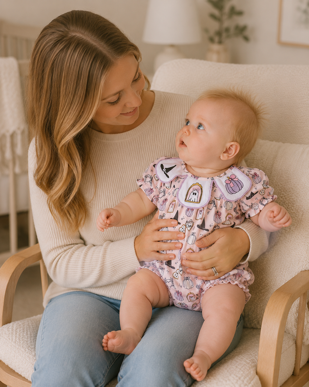 A baby wearing a pink and white patterned romper with a bib sits on a woman's lap in a cozy nursery chair. The woman is wearing a cream ribbed sweater and blue jeans.