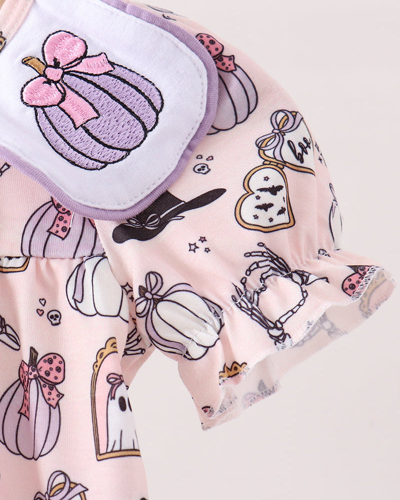 A close-up of a light pink short-sleeve top with a ruffled collar, featuring a whimsical print of pumpkins, ghosts, and stars. The collar is adorned with a white patch containing a purple embroidered pumpkin and a pink bow.