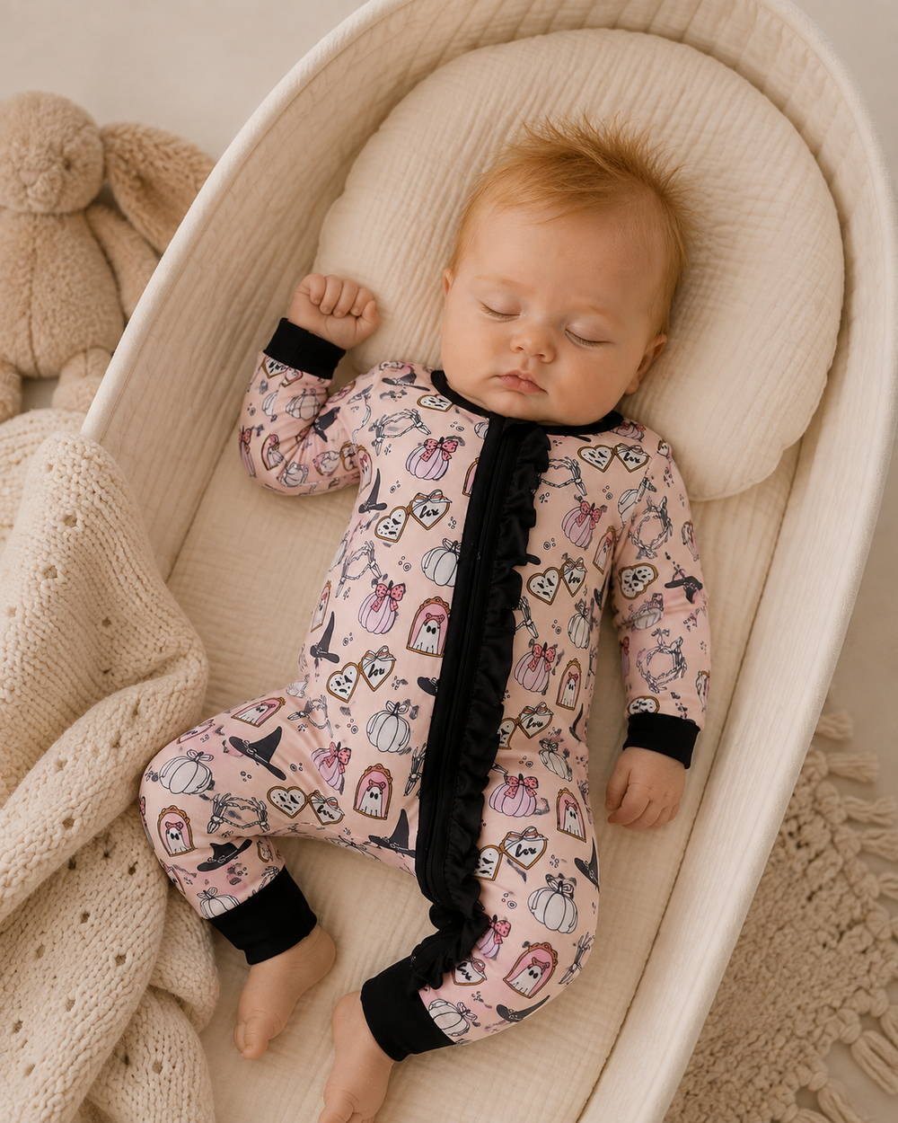 A sleeping baby wearing a pink full-body pajama with a black ruffled zipper and black cuffs, lying on a cream bassinet with a knitted blanket and a teddy bear nearby.